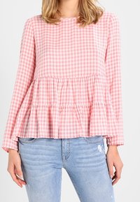 Donna che indossa una camicetta a maniche lunghe bianco e rosa a quadretti con orlo a peplo a più strati e jeans chiari strappati.