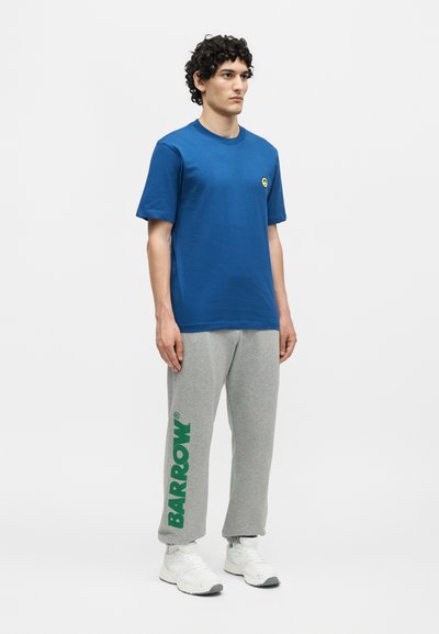 Άνδρας που στέκεται φορώντας μπλε μπλουζάκι με μικρό σήμα χαμογελαστής φατσούλας, γκρι φόρμα με πράσινο κείμενο "BARROW" και λευκά αθλητικά σε λευκό φόντο.