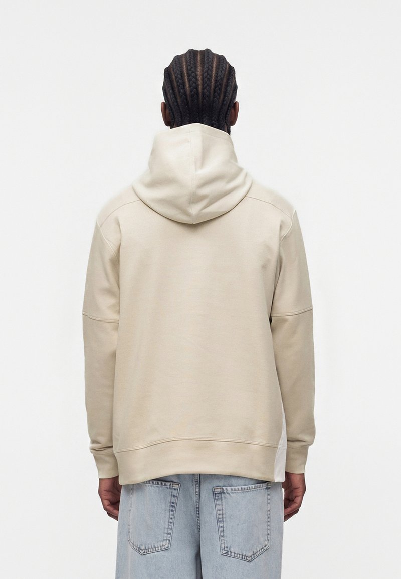 Mand med cornrow fletninger iført en beige hættetrøje og lyseblå jeans, stående med ryggen mod en ensfarvet hvid baggrund.