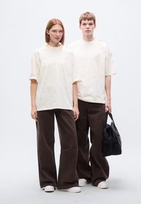 Δύο μοντέλα στέκονται μαζί, φορώντας ανοιχτόχρωμα κρεμ T-shirts με υφή και φαρδιά καφέ παντελόνια από κοτλέ, με μια μαύρη τσάντα στο χέρι του ανδρικού μοντέλου.