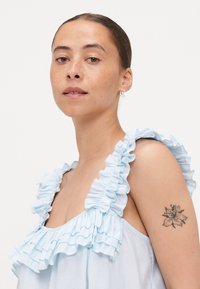 Lyseblå ermeløs bluse med volanger langs utringningen, laget av et mykt, teksturert stoff. En blomster-tatovering synlig på overarmen.