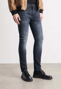 Jean skinny bleu foncé légèrement délavé, associé à des chaussures noires à lacets, présenté sur un fond gris clair.