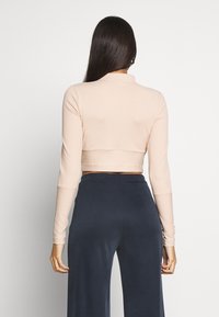 Långärmad, croppad topp i ljusbeige ribbad tyg med hög krage. Slät textur och åtsittande ärmar, i kombination med mörka byxor.