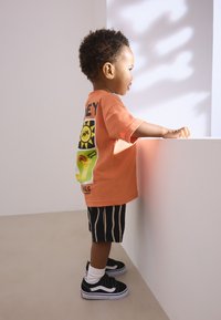Orange cotton t-shirt with a graphic on the back, black-striped shorts, and black and white sneakers with Velcro straps, standing by a white wall.