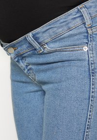 Jeans de denim azul claro com uma textura ligeiramente desbotada, apresentando um bolso para moedas na frente, metal em prata e costura laranja em contraste.