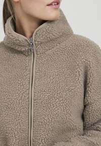 Beige fleecjas met een hoge kraag, textuur oppervlak en ritsluiting. Heeft een gladde binnenvoering en zilveren ritsbevestiging.
