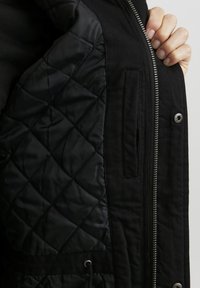 Černá bunda s prošívaným interiérem, zapínáním na zip, boční kapsou a stahovacími okraji. Exteriér má hladkou, matnou texturu.