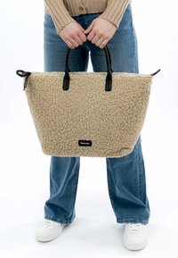 Beige textured tote bag with a fluffy material, black leather handles, and a small brand tag. The bag features a structured, wide shape.