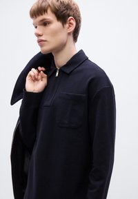 Μπλε σκούρο πουλόβερ με φερμουάρ στο γιακά, που διαθέτει τσέπη στο στήθος και μακριά μανίκια από απαλό, υφασμάτινο ύφασμα με υφή.