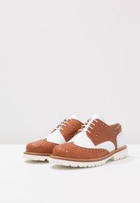 Chaussures en cuir marron et blanc style brogue à lacets, avec des détails perforés et des semelles épaisses en caoutchouc blanc sur une surface blanche.