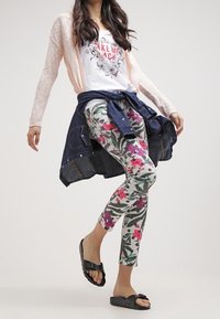 Mujer vestida con leggings florales, una camiseta gráfica blanca, un cárdigan rosa claro, sandalias negras y una chaqueta de mezclilla atada a la cintura, de pie con una pierna levantada.