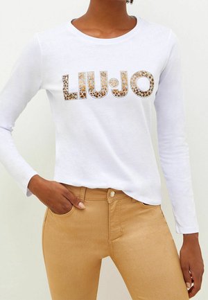 Person iført en hvid langærmet skjorte med gyldent paillet "LIU•JO" logo, kombineret med højtaljede beige bukser, med den ene hånd i lommen.