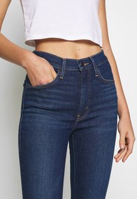 Vaqueros de talle alto azul oscuro con un diseño clásico, que cuentan con bolsillos delanteros, cremallera y detalles de costura dorados. Textura de denim suave.