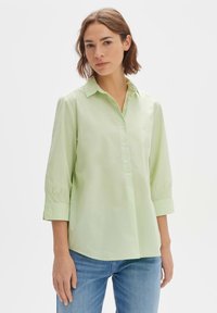 Camicia verde chiaro con colletto, chiusura a bottoni e maniche a tre quarti. Il tessuto appare liscio, con dettagli arricciati alla fine delle maniche.