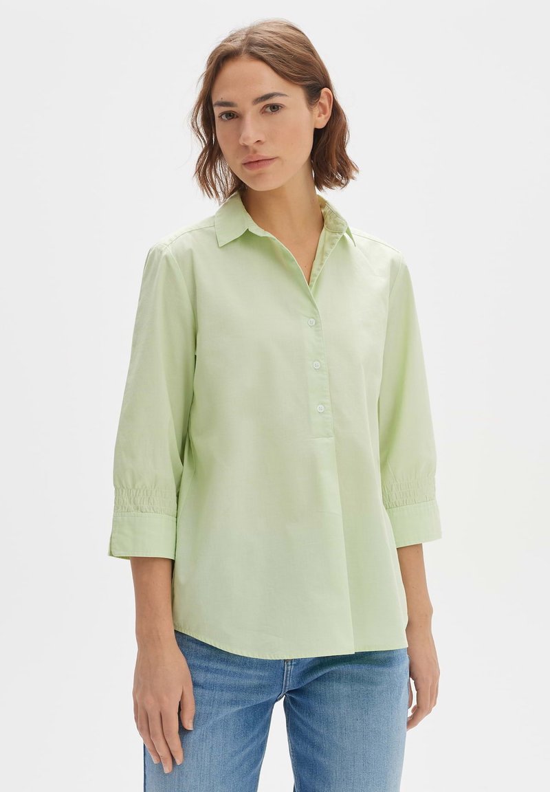 Camicia verde chiaro con colletto, chiusura a bottoni e maniche a tre quarti. Il tessuto appare liscio, con dettagli arricciati alla fine delle maniche.