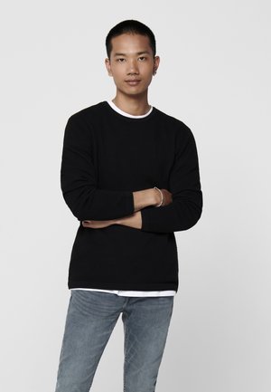 Black textured sweatshirt with a round neckline, layered over a white shirt, paired with light blue jeans. Model stands with arms crossed.