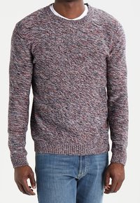 Pull à col rond en maille texturée avec un mélange de fibres grises, rouges et blanches. Poignets et ourlet ajustés, porté par-dessus un T-shirt blanc.