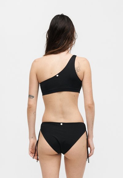 Γυναίκα με μακριά καστανά μαλλιά φοράει μαύρο μπικίνι με έναν ώμο και μαύρα κάτω μέρος μπικίνι με δεσίματα στα πλάγια, φανερώνεται από πίσω.