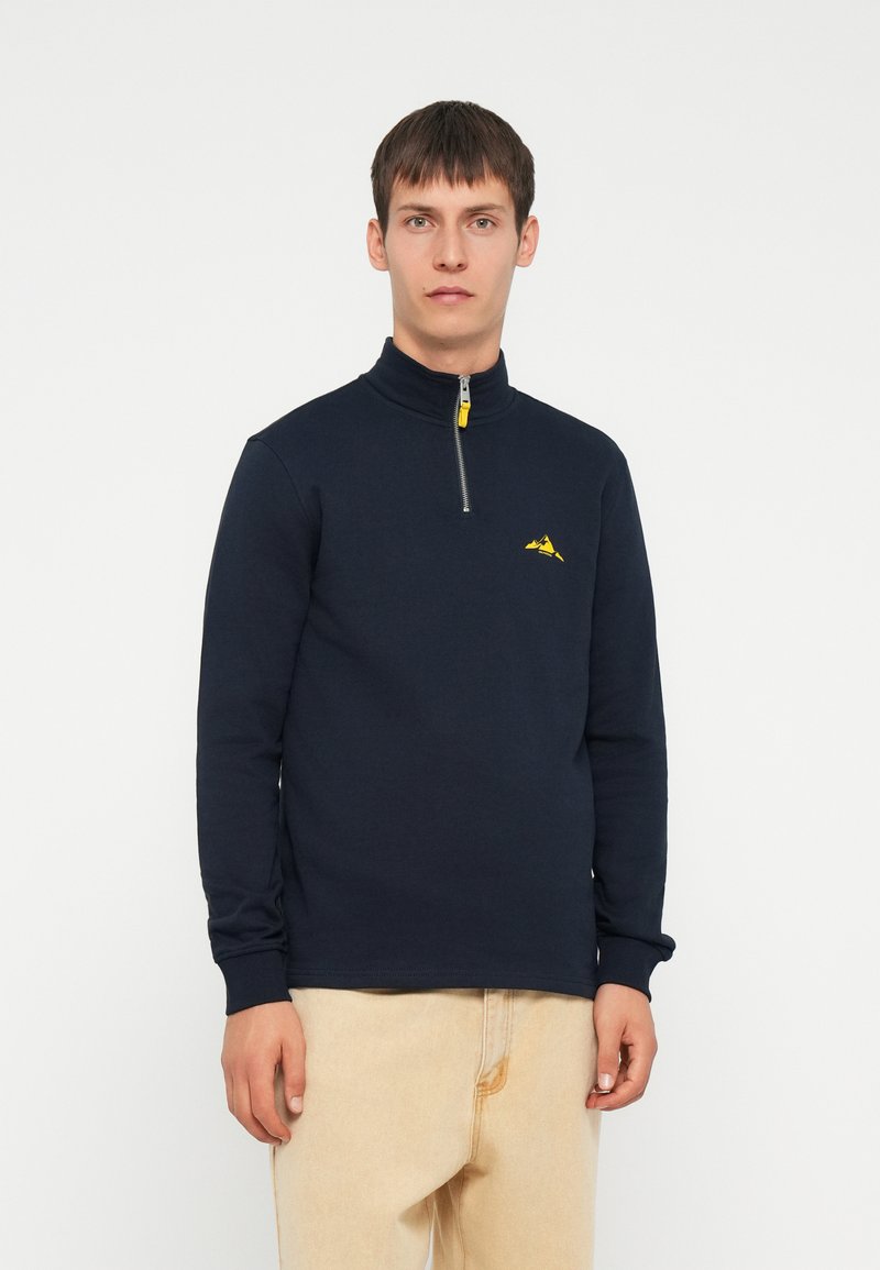Marineblauwe sweatshirt met rits en hoge kraag, met een gele rits en een klein geel berglogo op de borst. Gladde textuur.