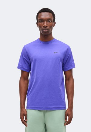 Férfi, aki lila Nike rövid ujjú pólót visel, a mellkasán kis fekete logóval, világoszöld rövidnadrággal párosítva, sima háttér előtt áll.