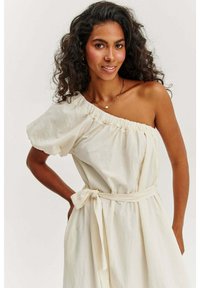 Krémové šaty na jedno rameno vyrobené z jemného materiálu s naberaným výstrihom a zviazaným pásom, ktoré predvádzajú uvoľnenú siluetu.