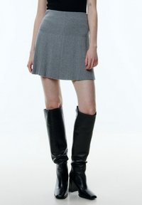 Femme portant une mini-jupe plissée grise, un top sans manches noir et des bottes en cuir noires à talons hauts jusqu'aux genoux.