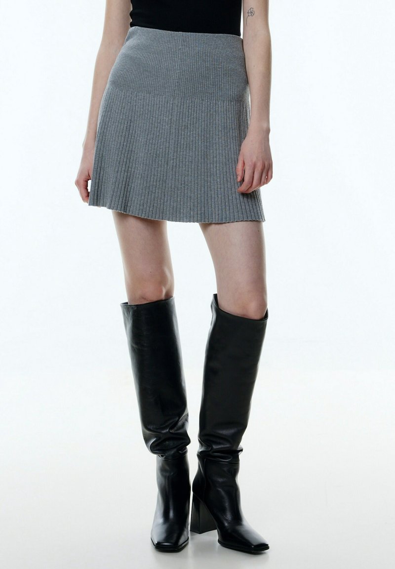 Femme portant une mini-jupe plissée grise, un top sans manches noir et des bottes en cuir noires à talons hauts jusqu'aux genoux.