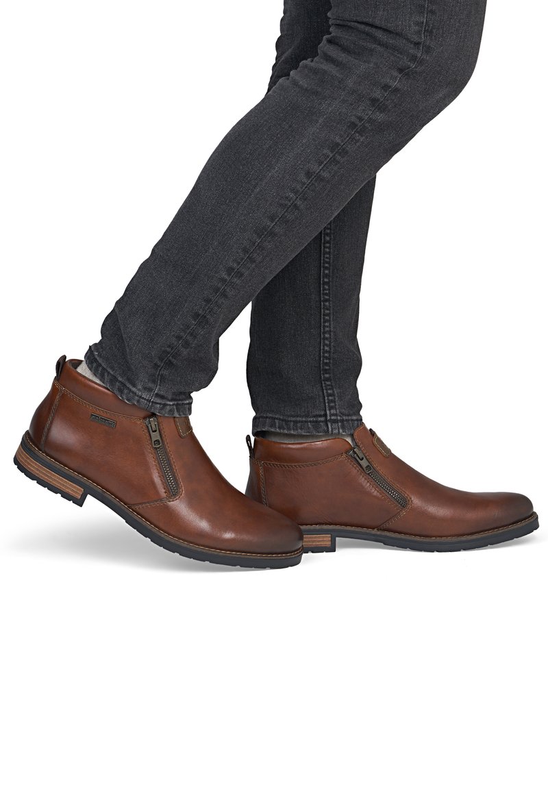 Bottines en cuir marron avec fermetures éclair latérales portées avec un jean noir slim, montrées en position de marche sur fond blanc.