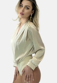 Beige silke bodystocking med lange ærmer, draperet front og tætsiddende design; har en glat tekstur og delikate manchetter. Guld hoops øreringe.