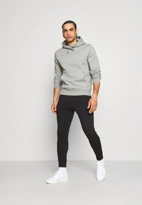 Siv pulover s kapuco z vrvico, kombiniran s črnimi zoženimi hlačami. Model nosi bele superge in stoji na nevtralnem ozadju.