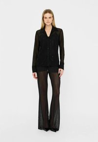 Mustast läbipaistvast siidist pluus, millel on pikad varrukad ja nööbid, kombineeritud sobivate lehvikukujuliste pükstega. Mõlemal on dekoratiivne muster ja voogav tekstuur.