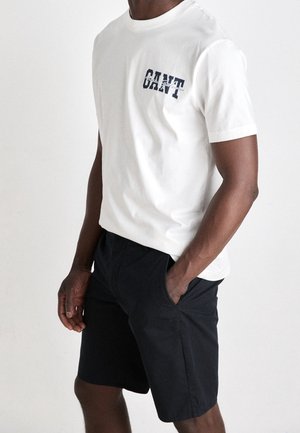 Biała bawełniana koszulka z granatowymi grafikami z napisem „GANT”. Połączona z czarnymi szortami, prezentująca luźny, swobodny krój i gładką fakturę.