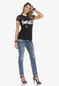 Schwarzes T-Shirt mit Mesh-Halsausschnitt und weißem Text "ANGEL" im Splatter-Paint-Design. Kombiniert mit blauen Skinny Jeans und schwarzen Absatzsandalen.