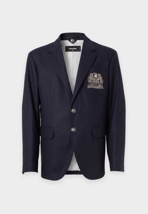 Homme portant un blazer bleu marine, une chemise blanche, un pantalon beige, une casquette marron et des bottes noires, tenant un sac en cuir noir, regardant sur le côté avec une expression neutre.