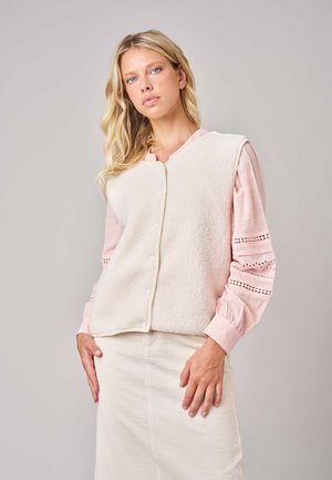 Vrouw met lang blond haar draagt een crèmekleurig fleece gilet met knopen over een roze blouse met kanten mouwen en een crèmekleurige geribbelde rok.