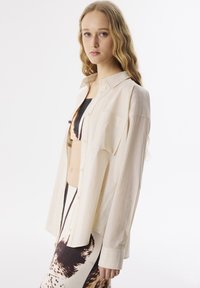 Kremowa koszula z długim rękawem wykonana z lekkiego, teksturowanego materiału. Jedna kieszeń na piersi; noszona na wzorzystą maxi spódnicę.