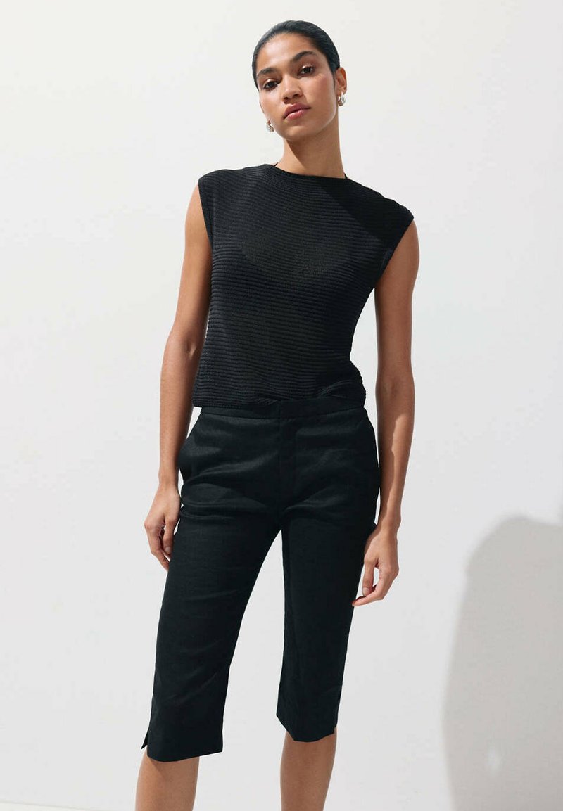 Femme portant un haut noir sans manches et un pantalon noir au genou, debout devant un fond clair uni avec une main détendue.