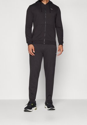 Homme portant un sweat à capuche noir zippé et un pantalon de jogging noir assorti, avec des chaussures de sport noires et blanches sur fond blanc.