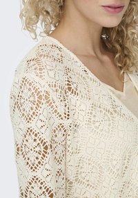 Cardigan in pizzo di colore crema con intricati motivi floreali, maniche lunghe e scollatura a V profonda, che mette in mostra un design leggero e testurizzato.