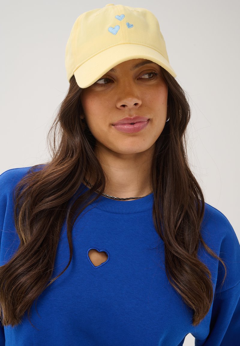 Femme aux longs cheveux foncés portant une casquette jaune avec des cœurs bleus et un sweat-shirt bleu avec une découpe en forme de cœur près de la clavicule.