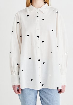 Hvid bluse med lange ærmer med flæset krave og sorte hjerte mønstre, båret uden på lyseblå jeans.