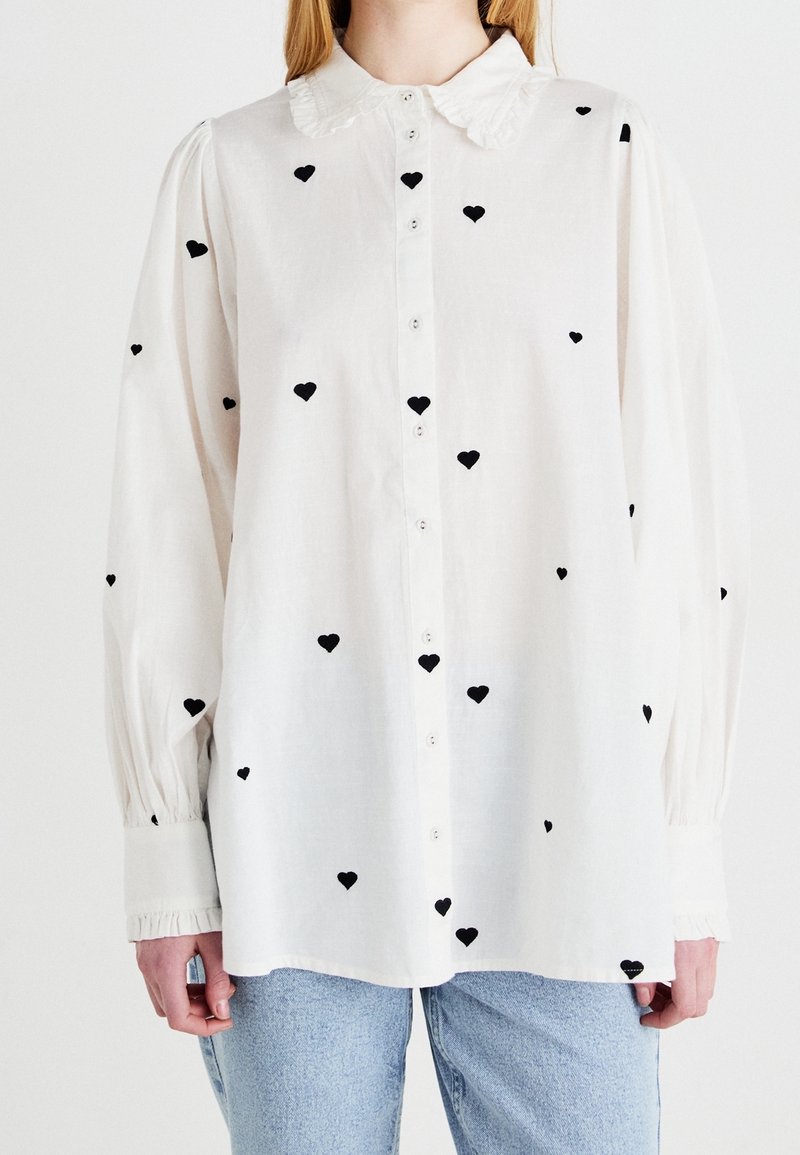 Blouse blanche à manches longues avec col à volants et motifs de cœurs noirs, portée rentrée sur un jean bleu clair.