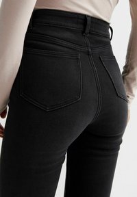 Jeans de denim negro de cintura alta con una textura suave, costuras en contraste y dos bolsillos traseros. Modelo visto desde la parte trasera.