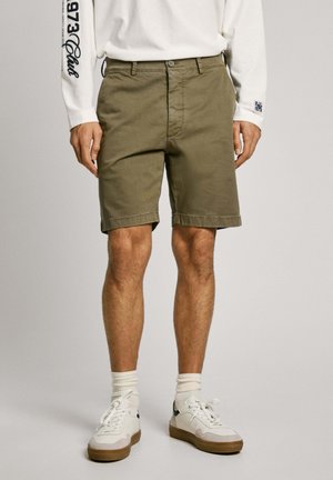 Olijfgroene katoen shorts met een rechte snit, voor bediening met knoopsluiting en omgeslagen zoom. Gekleed met witte sneakers en gestreepte sokken.