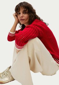 Roter, gestrickter Pullover mit gemusterter Textur und weißen gestreiften Bündchen. Kombiniert mit creamfarbenen, weit geschnittenen Hosen und goldenen Sportschuhen.