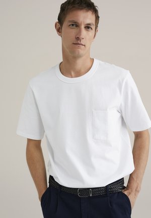Camiseta de algodón blanca con mangas cortas, escote redondo y un bolsillo en el pecho izquierdo. Llevada con un cinturón trenzado negro sobre unos pantalones azul marino.