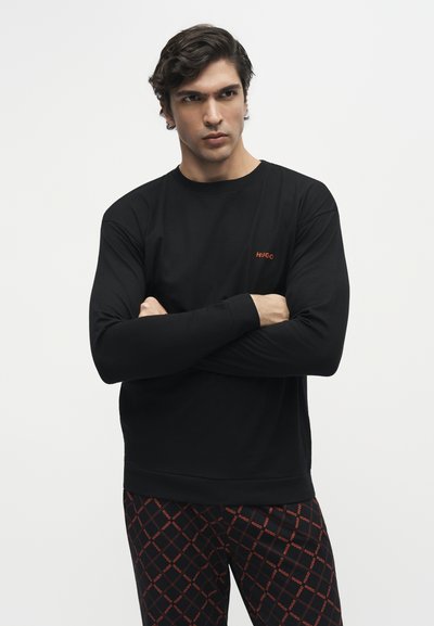 Jeune homme les bras croisés portant un t-shirt noir à manches longues et un pantalon noir avec un texte rouge à motifs sur un fond blanc uni.