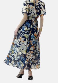 Navy sinivuokkoja kuviollinen pitkä mekko, jossa on läpinäkyvä kangas, puffihihat, smokkikaulus ja monivärinen kukkakuvaus. Mustat nauhalliset korkokengät.