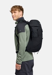 Sac à dos noir avec plusieurs compartiments, éléments texturés, sangles ajustables et une forme épurée, porté sur une chemise verte à manches longues.