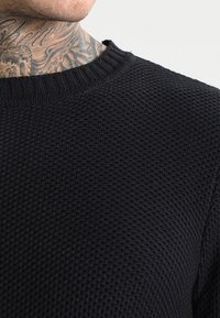 Svart strukturerad stickad tröja med ribbad krage och en kroppsnära passform som betonar ett vävt mönster genom hela tyget.